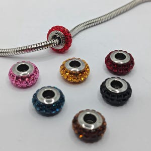 May include: Seven charm beads with a silver metal base and a variety of colored crystals. The beads are red, pink, orange, blue, black, and two shades of brown.