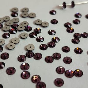 May include: A collection of purple rhinestones and silver-colored round sequins. The rhinestones are scattered on a white surface.