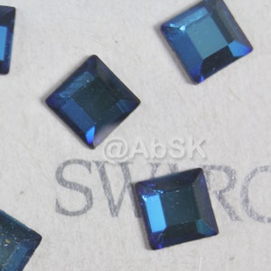 May include: Close-up of several square-shaped blue rhinestones on a white surface with the word "SWAROVSKI" printed in grey.