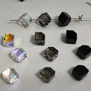 Könnte beinhalten: Sortiment an facettierten Glasperlen in verschiedenen Farben, darunter klar, schillernd, schwarz und grau. Die Perlen sind würfelförmig und haben eine glänzende Oberfläche.