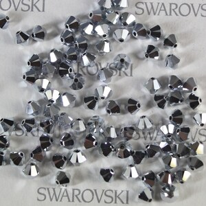 Op de afbeelding: Een close-up van een stapel zilverkleurige, gefacetteerde kristallen kralen. De kralen zijn verspreid over een wit oppervlak met het woord "SWAROVSKI" erop gedrukt.