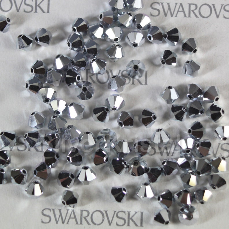 Op de afbeelding: Een close-up van een stapel zilverkleurige, gefacetteerde kristallen kralen. De kralen zijn verspreid over een wit oppervlak met het woord "SWAROVSKI" erop gedrukt.