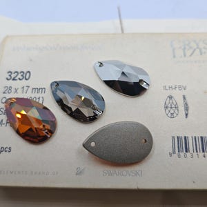 Op de afbeelding: Een close-up van een verpakking met 24 Swarovski-kristallen in druppelvorm. De druppels zijn in drie kleuren: amber, zwart en zilver. De verpakking bevat het productnummer 3230, de maat 28 x 17 mm en de tekst "THE ELEMENTS BRAND OF SWAROVSKI".