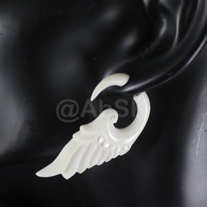 Puede incluir: Pendiente de hueso blanco en forma de ala con un gancho curvo. El pendiente se lleva en la oreja de una persona.