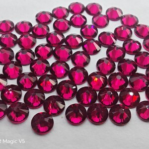 Op de afbeelding: Een verzameling van talloze kleine, ronde, gefacetteerde fuchsia-kleurige kristallen gerangschikt in een losse groep. Elk kristal heeft een puntige bovenkant en een reflecterend oppervlak dat het licht vangt. De tekst HONOR Magic V5 is zichtbaar.
