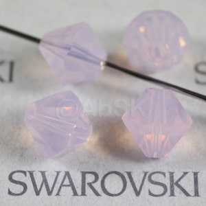May include: Four light purple faceted glass beads with a silver wire running through them. The beads are on a white surface with the word "SWAROVSKI" printed on it.