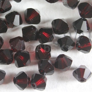 May include: A group of dark red crystal beads with a faceted design. The beads are scattered on a white surface.