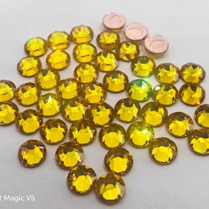 Può includere: Una collezione di strass sfaccettati, rotondi, giallo-oro e alcuni piccoli dischi metallici color rame. Gli strass sono sparsi su una superficie bianca, catturando la luce e mostrando una brillante lucentezza. L'immagine è ben illuminata, evidenziando le gemme.