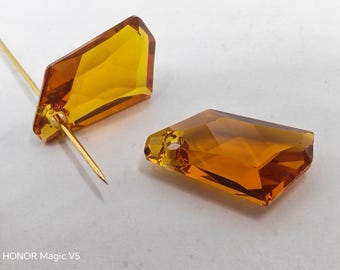 May include: Two amber-colored, faceted crystal beads. One bead is attached to a gold-colored pin, while the other lies separately. The crystals have a geometric, rectangular shape with a hole for stringing. The image is taken on a white surface.