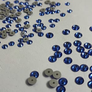 May include: A scattering of blue iridescent sequins on a white surface. The sequins are small and round, with a flat back and a raised, shiny front.