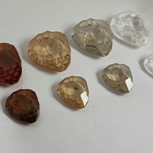 May include: A selection of faceted, teardrop-shaped beads in shades of brown, amber, and clear. The beads are holed at the top for stringing and are arranged on a white surface. The facets catch the light, creating highlights.