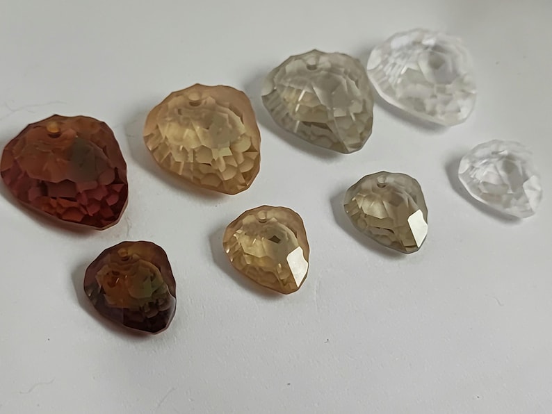 May include: A selection of faceted, teardrop-shaped beads in shades of brown, amber, and clear. The beads are holed at the top for stringing and are arranged on a white surface. The facets catch the light, creating highlights.