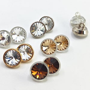 May include: A selection of round crystal buttons in various colours, including clear, gold, and brown. The buttons have a faceted design and are set in silver or gold-toned metal settings. Some buttons have a clear plastic backing.