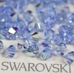 Peut inclure: Gros plan sur un tas de perles en cristal bleu clair avec un reflet arc-en-ciel iridescent. Les perles sont facett&eacute;es et ont un trou au centre. Les perles sont dispos&eacute;es sur une surface blanche avec le mot "SWAROVSKI" imprim&eacute; en noir.