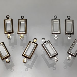 May include: Eight clear glass rectangular settings with gold and silver metal frames. The settings have two loops on the top for attaching to a necklace or bracelet.