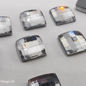 May include: Close-up of several clear, square-shaped rhinestones with faceted surfaces. The rhinestones are arranged on a light gray surface, reflecting light and creating a sparkling effect. The image is well-lit, highlighting the details of the crystals.