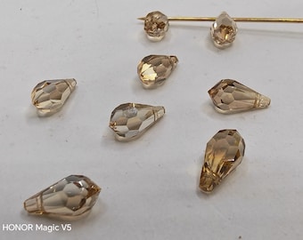 May include: Close-up of several faceted, teardrop-shaped beads in shades of amber and gold. A gold-colored pin with two beads is also visible. The beads are scattered on a light-colored surface, ideal for jewelry making or crafting.