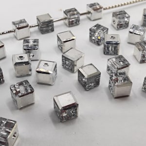 Op de afbeelding: Close-up van heldere, kubusvormige kralen met zilverkleurige metalen accenten. Sommige kralen zijn geregen op een zilveren draad, andere zijn verspreid over een wit oppervlak. Ideaal voor het maken van sieraden.