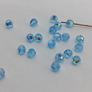 Puede incluir: Un conjunto de cuentas facetadas de color azul claro dispersas sobre una superficie blanca. Las cuentas tienen una cualidad iridiscente y brillante. También hay un fino alambre de color cobre, con una cuenta ensartada. Estas cuentas son adecuadas para la fabricación de joyas.