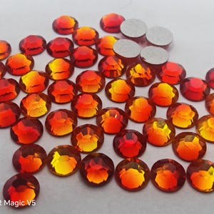 Könnte beinhalten: Eine Ansammlung runder, facettierter orangefarbener und roter Kristalle mit reflektierender Oberfläche. Einige Kristalle liegen mit der flachen, silberfarbenen Kleberückseite nach oben, während die anderen ihre leuchtenden, mehrfarbigen Oberflächen zeigen.