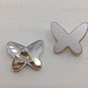 Pode incluir: Dois enfeites em forma de borboleta, um de cristal transparente e outro prateado, sobre um fundo branco com a palavra "SWAROVSKI" impressa.