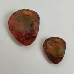 May include: Two faceted, teardrop-shaped pendants. The larger pendant is a deep reddish-brown with hints of green and yellow. The smaller pendant has similar colouring. Both are set against a white background.