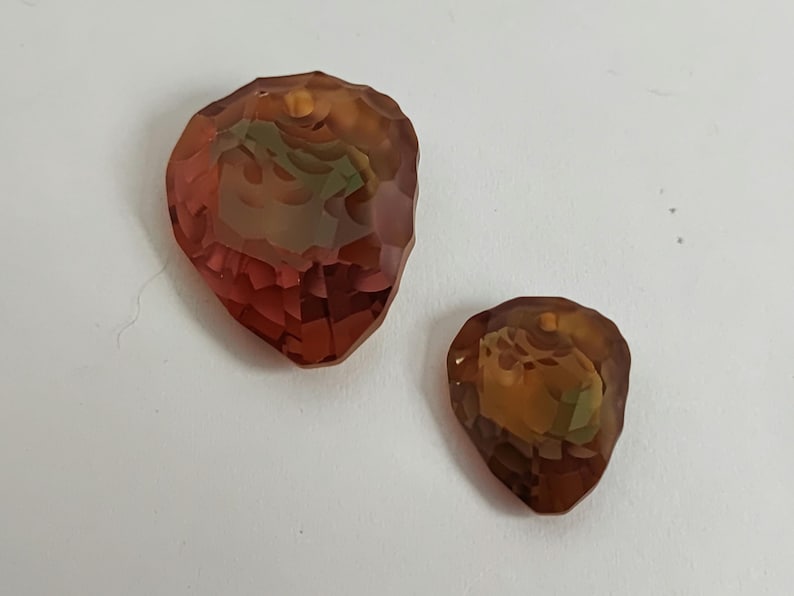 May include: Two faceted, teardrop-shaped pendants. The larger pendant is a deep reddish-brown with hints of green and yellow. The smaller pendant has similar colouring. Both are set against a white background.