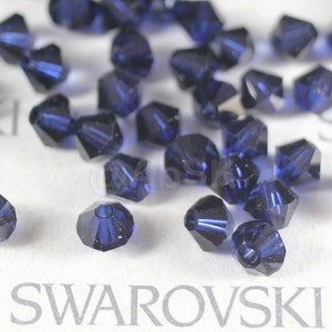 May include: A close-up of a group of dark blue crystal beads. The beads are faceted and have a pointed shape. The beads are scattered on a white surface with the word "SWAROVSKI" printed on it.