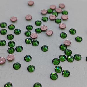 May include: Close-up of scattered green and pink rhinestones. The green rhinestones are round with a faceted surface, reflecting light. The pink rhinestones are also round, with a flat, metallic appearance. The rhinestones are scattered on a light-colored surface.