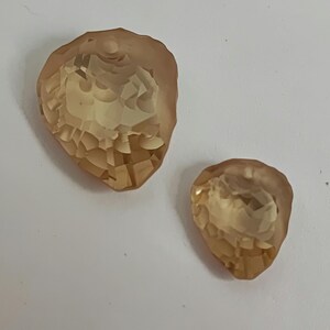 May include: Two amber and peach-coloured, faceted, teardrop-shaped crystals. The crystals have a textured, geometric pattern and a hole at the top for stringing. They are set against a white background.