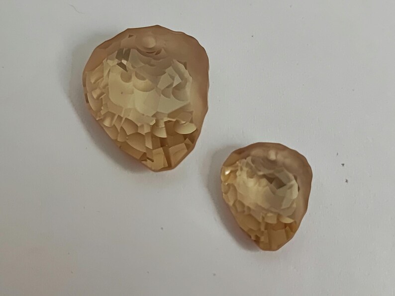 May include: Two amber and peach-coloured, faceted, teardrop-shaped crystals. The crystals have a textured, geometric pattern and a hole at the top for stringing. They are set against a white background.