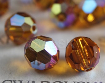 Puede incluir: Primer plano de un grupo de cuentas de vidrio ámbar iridiscente. Las cuentas son facetadas y tienen un acabado brillante. Las cuentas están dispuestas sobre una superficie blanca con la palabra "SWAROVSKI" impresa en negro.