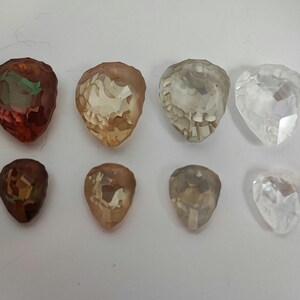 May include: Eight faceted teardrop-shaped gemstones in shades of brown, amber, and clear. The gemstones are arranged in two rows of four, with the top row featuring larger stones. The stones have a polished, reflective surface.