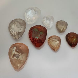 May include: A collection of faceted gemstone beads in shades of clear, red, and peach. The teardrop-shaped beads are displayed on a white surface, highlighting their reflective properties and intricate cuts. Measurements are in cm.