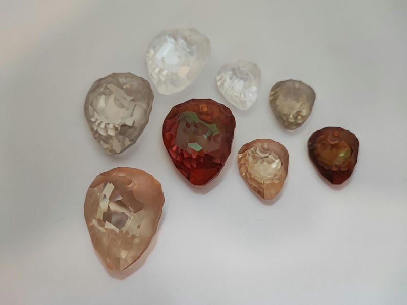 May include: A collection of faceted gemstone beads in shades of clear, red, and peach. The teardrop-shaped beads are displayed on a white surface, highlighting their reflective properties and intricate cuts. Measurements are in cm.