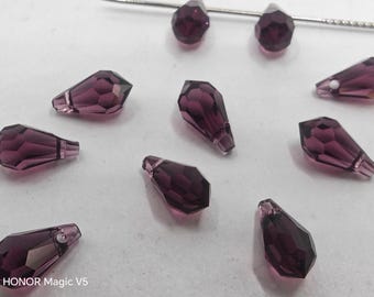 May include: A collection of faceted, teardrop-shaped beads in a deep purple hue. The beads are arranged on a white surface, with a silver needle visible in the background. These beads are likely for jewelry making or other craft projects.