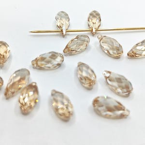 Op de afbeelding: Een verzameling gefacetteerde, druppelvormige kralen in een warme amberkleur. De kralen liggen verspreid op een witte ondergrond, met een goudkleurige speld die door twee ervan gaat. Geschikt voor het maken van sieraden.