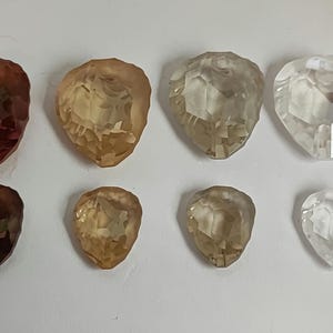 May include: Eight faceted gemstones in shades of brown and clear. The teardrop-shaped gems are arranged in two rows of four. The colours range from deep reddish-brown to pale yellow and clear.