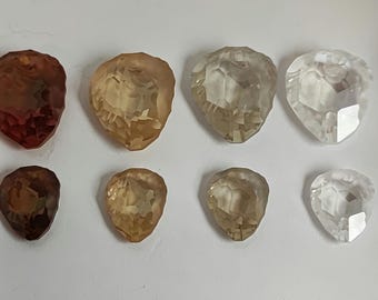 May include: Eight faceted gemstones in various shades of brown and clear. The gemstones are teardrop-shaped and arranged in two rows of four. The colors range from deep reddish-brown to pale yellow and clear.