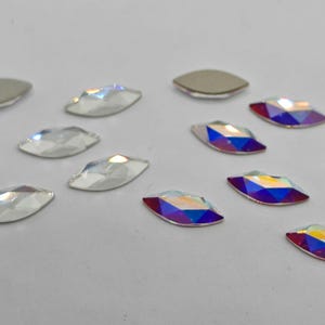 May include: Assortment of clear and iridescent glass rhinestones in a marquise shape. The rhinestones are arranged in two rows on a white surface.