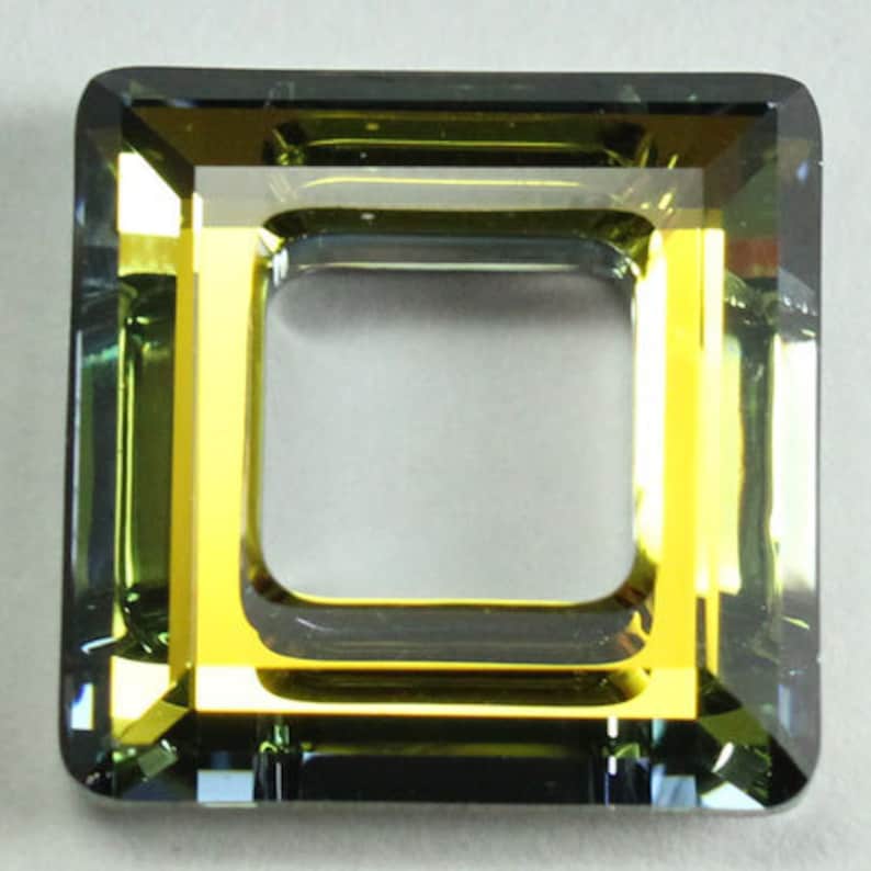 Puede incluir: Una cuenta de vidrio de forma cuadrada, facetada, con un marco interior de color dorado. La cuenta es de color verde oscuro con un ligero tono azul.