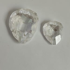 May include: Two clear, faceted crystal teardrop-shaped pendants. The larger pendant is on the left, and the smaller one is on the right. Both have multiple facets that reflect light, creating a sparkling effect. The pendants are set against a plain white background.