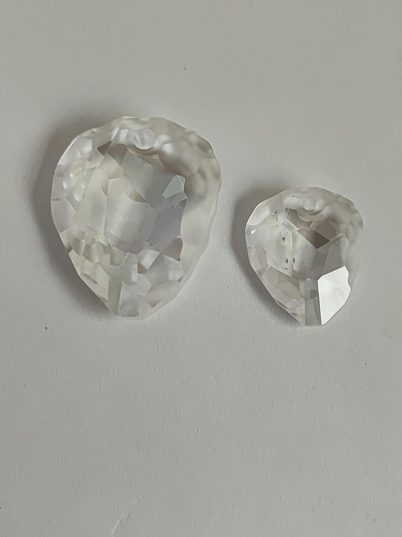 May include: Two clear, faceted crystal teardrop-shaped pendants. The larger pendant is on the left, and the smaller one is on the right. Both have multiple facets that reflect light, creating a sparkling effect. The pendants are set against a plain white background.
