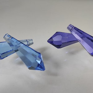 May include: Two crystal prisms, one blue and one purple, are shown on a white surface. The prisms are faceted and have a pointed tip.