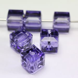 May include: A group of six purple square glass beads with a hole in the center. The beads are faceted and have a clear center.