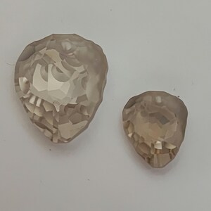 May include: Two faceted, teardrop-shaped crystals in shades of light brown. The larger crystal is approximately 3.8 cm long, while the smaller one is about 2.5 cm. Both have multiple facets, reflecting light.