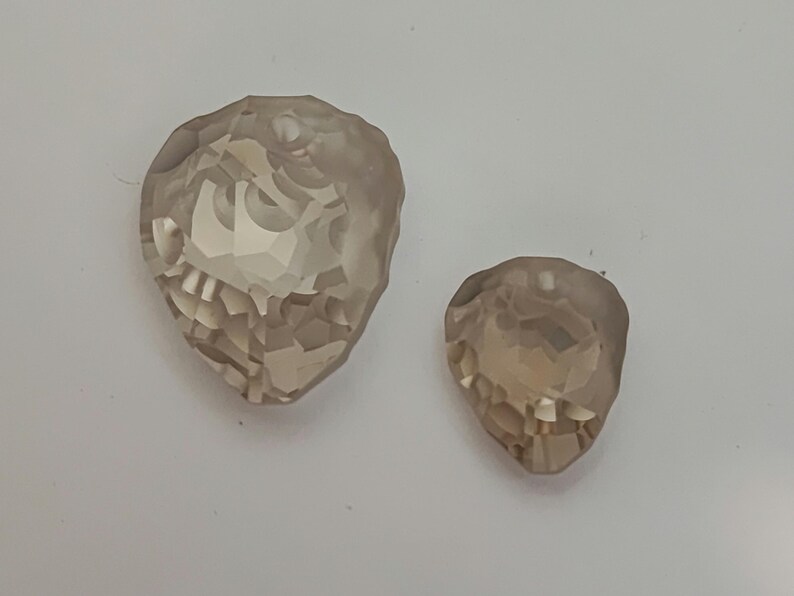 May include: Two faceted, teardrop-shaped crystals in shades of light brown. The larger crystal is approximately 3.8 cm long, while the smaller one is about 2.5 cm. Both have multiple facets, reflecting light.
