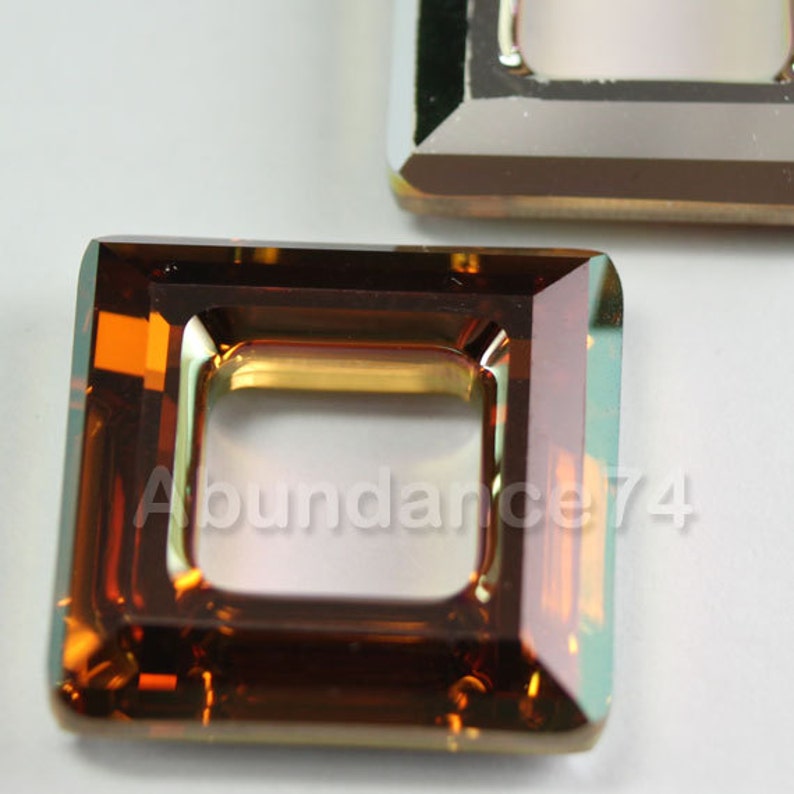 May include: Close-up of two square-shaped, faceted glass beads. The beads are a deep amber colour with a square cut-out in the centre. The text "Abundance74" is visible on the bottom bead. The edges of the beads have a slight iridescent sheen.