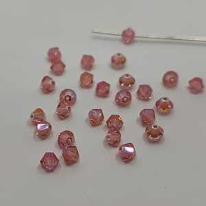 Puede incluir: Una colección de cuentas de cristal rosa con un diseño facetado. Las cuentas están esparcidas sobre una superficie blanca.
