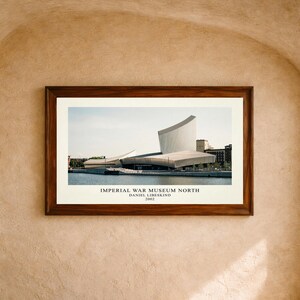 May include: A framed architectural print of the Imperial War Museum North by Daniel Libeskind, 2002. The image features the angular, metallic-toned building situated along a waterfront, presented in a dark wood frame with a white mat border.
